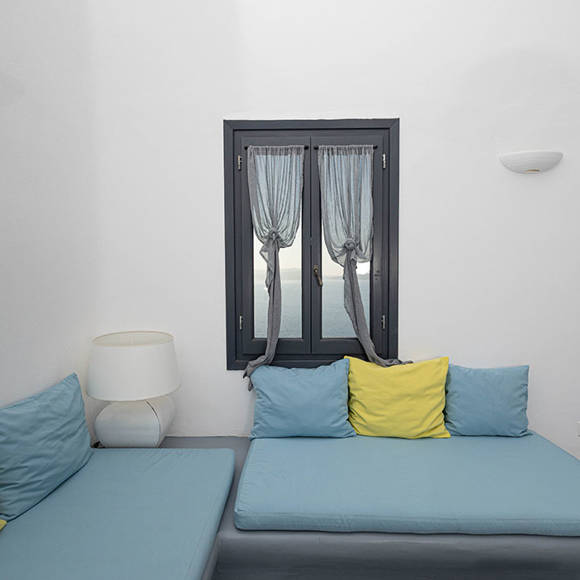 Grand Ambassador Santorini Serenity Two Bedroom Suite living area with twin built-in blue sofas, yellow accent pillow, window with caldera view