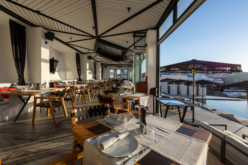 Grand Ambassador Dione restaurant interior with wooden chairs, set tables, and view of pool and umbrellas