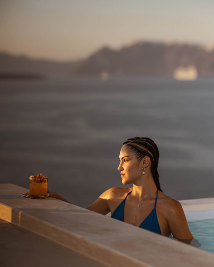 Guest relaxing in infinity pool with drink at Grand Ambassador hotel overlooking caldera views