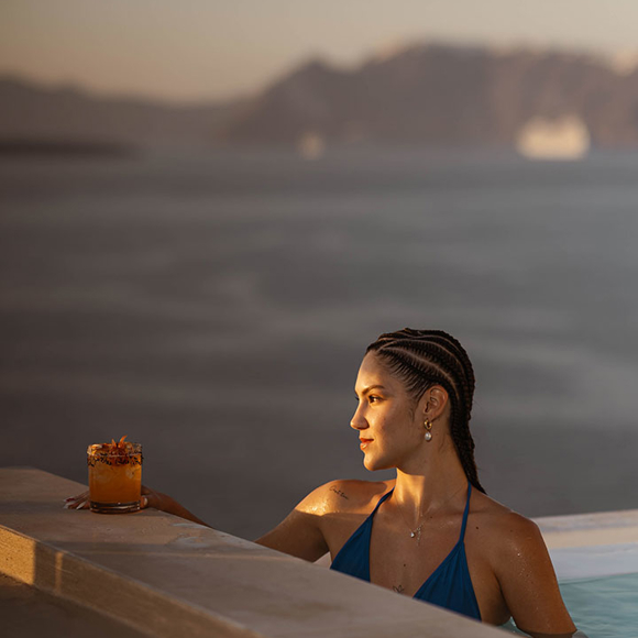 Guest relaxing in infinity pool with drink at Grand Ambassador hotel overlooking caldera views