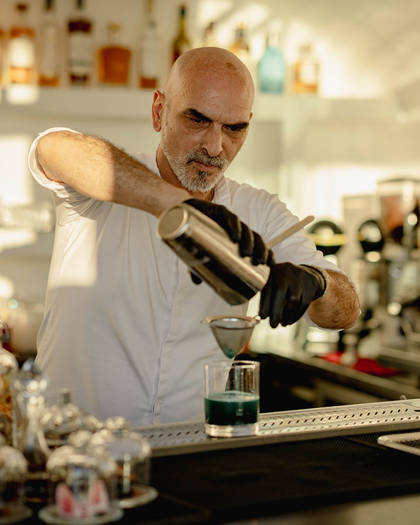 Grand Ambassador pool bar bartender wearing gloves pouring blue cocktail through strainer into glass with bottles in background